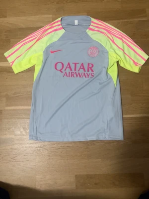 PSG Nike matchtröja ljusgrå/rosa - Snygg PSG matchtröja från Nike i ljusgrått med neon-gula och rosa detaljer. Tröjan har korta ärmar med rosa ränder och PSG-logga samt Nike Swoosh på bröstet. Tillverkad i lätt och ventilerande funktionsmaterial, perfekt för träning eller match.
