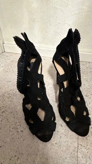 Svarta cut out pumps med klack - Säljer ett par svarta pumps med hög klack och unika cut out-detaljer. Skorna har en öppen design med dekorativa kanter och dragkedja bak. Materialet ser ut att vara mocka eller mockaimitation. Perfekta för dig som vill sticka ut med din stil.