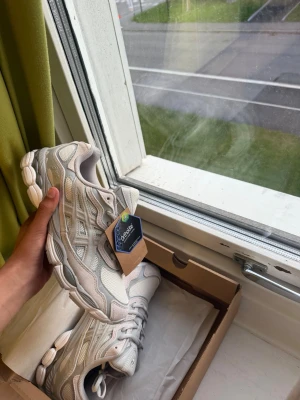 Asics gel nyc cream/cloud gray 41,5 - Snygga vita ASICS sneakers med silvriga detaljer och klassisk logga på sidan. Skorna har en sportig design med meshpaneler för extra andningsförmåga och en chunky sula som ger skön komfort. Perfekta för dig som gillar stilrena och trendiga sneakers.