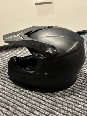 Svart crosshjälm XS - Säljer en svart crosshjälm i storlek XS med skärm och ventiler. Hjälmen har röd och svart vadderad insida för extra komfort och skydd. Passar perfekt för motocross eller liknande aktiviteter där du behöver extra säkerhet.