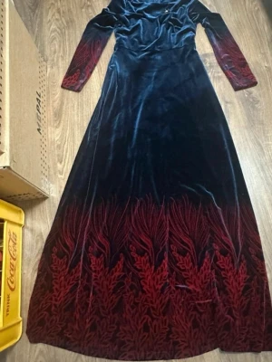 Lång sammetsklänning i mörkblått och rött - Elegant långklänning i mörkblå sammet med röda broderade detaljer längs ärmar och nederkant. Klänningen har lång ärm och figurnära passform upptill, med utsvängd kjol. Perfekt för dig som vill sticka ut med en unik och lyxig look.