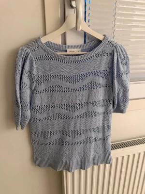 Ljusblå stickad topp från Vila - Söt ljusblå stickad topp från Vila med kort puffärm och vågigt hålmönster. Rund halsringning och ribbade kanter. Perfekt för dig som gillar en feminin och trendig stil. Materialet känns mjukt och luftigt.