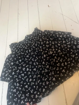 Svart blommig volangkjol - Supersöt svart kortkjol med vita blommor och volangdetaljer. Kjolen har smock i midjan för en bekväm passform och är perfekt för sommarens alla äventyr. Materialet känns lätt och luftigt, vilket gör den enkel att styla med en enkel topp. lite nopprig men annars i ett helt okej skick. Inga hål eller fläckar.