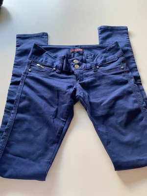 Marinblå stretchbyxor - Snygga marinblå jeans med dubbla silverknappar och klassisk femficksdesign. Jeansen har raka ben och detaljerade sömmar samt små metallnitar vid fickorna. Materialet är mjukt och stretchigt för extra komfort.