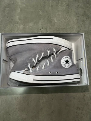 Grå Converse Chuck Taylor Highs - Säljer ett par grå Converse Chuck Taylor All Star high tops med vit tåhätta, vita snören och klassisk svart rand runt sulan. Skorna är i canvasmaterial och har den ikoniska loggan på sidan. Perfekta för en avslappnad och stilren look.