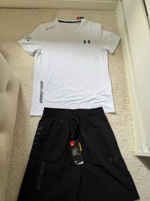 Under Armour träningsset svart/vit - Snyggt träningsset från Under Armour med en vit t-shirt och svarta shorts. T-shirten har tryckt logga på bröstet och text på sidan, medan shortsen har elastisk midja med dragsko och logga på benet. Perfekt för gymmet eller löpning.