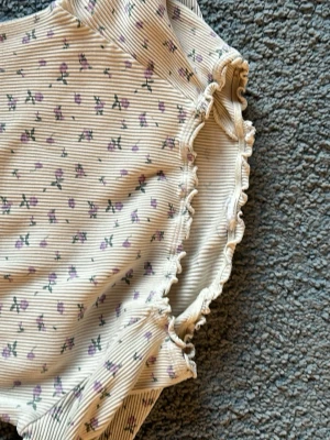 Blommig ribbad långärmad topp - Supersöt långärmad topp i ljusbeige med små lila blommor och tunna ränder. Ribbad struktur och volangdetaljer vid ärmslut, halsringning och nederkant. Perfekt för dig som gillar romantisk och trendig stil.