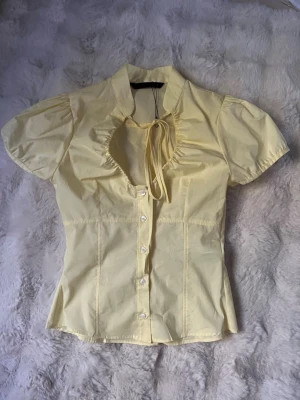 Gul blus med knappar och rosett - Söt ljusgul blus med korta puffärmar, knappar framtill och en dekorativ rosett vid halsringningen. Figurnära passform och lätt rynkade detaljer vid halsen ger en feminin look. Perfekt till jeans eller kjol.