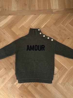 Grön stickad tröja från Zadig och Voltaire  - Mörkgrön stickad tröja med hög krage och fyra vita knappar längs axeln. Svart text 'AMOUR' på bröstet. Tröjan har ribbstickade muddar och är gjord i ett mjukt material, perfekt för att hålla sig varm. Långärmad och avslappnad passform 🤗❤️. Säljer för att den inte längre passar. 
