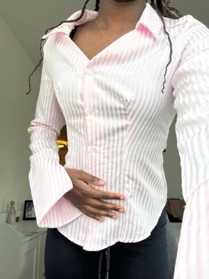 Figursydd skjorta med vida ärmar - Säljer en ljusrosa skjorta med vita ränder och vida armar med slits i storlek xs. Skjortan har klassisk krage, knappar framtill och figurnära passform. Perfekt för dig som gillar en feminin och trendig look. Aldrig använt.