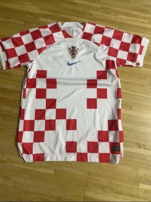 Kroatien Nike fotbollströja röd/vit - Säljer en officiell Kroatien fotbollströja från Nike i vitt med röda schackrutor och blå Nike-logga. Tröjan har Kroatiens landslagsemblem på bröstet och är tillverkad i lätt, ventilerande material. Perfekt för match eller träning.