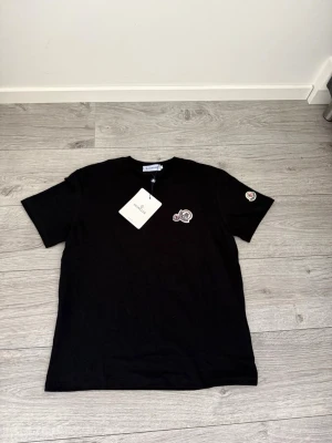 Svart Moncler t-shirt storlek M - Snygg svart t-shirt från Moncler med klassisk logga på bröstet och ärmen. T-shirten har rund halsringning och korta ärmar. Tillverkad i mjuk bomull som känns skön mot huden. Perfekt för en clean och stilren look. Kom dm för frågor, bud eller byten