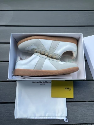 Maison Margiela vita sneakers - Maison Margiela sneakers i vitt läder och ljusgrå mockadetaljer. Klassisk låg modell med beige snören och gummisula i brunt. Snygg minimalistisk design som passar till det mesta. Kommer med originalkartong och dustbag.