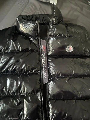 Svart dunväst från Moncler - Snygg svart dunväst från Moncler med glansig finish och hög krage. Västen har Moncler-logga på bröstet och tydlig Moncler-text på dragkedjans kantband. Perfekt för lager-på-lager och streetwear-stil.