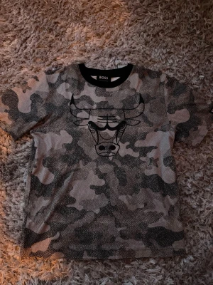 Camouflage Chicago Bulls t-shirt BOSS - Snygg t-shirt från BOSS x NBA med Chicago Bulls-tryck framtill. T-shirten har ett grått och svart camouflage-mönster, rund halsringning med svart kant och är kortärmad. Perfekt för dig som gillar streetwear och basket.