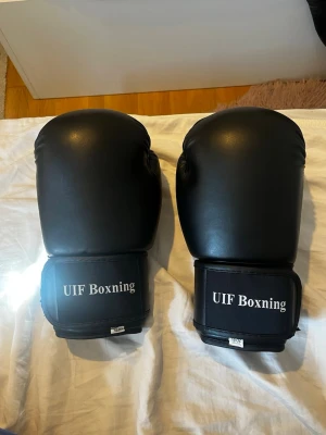 Svarta boxningshandskar UIF Boxning - Ett par svarta boxningshandskar från UIF Boxning i konstläder med kardborreknäppning runt handleden. Handskarna har en klassisk form och är vadderade för bra skydd vid träning eller sparring.
