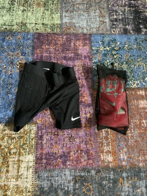 Nike målvaktsbyxor och handskar - Säljer ett par svarta målvaktsbyxor från Nike med vadderade sidor för extra skydd samt ett par målvaktshandskar i rött och vitt. Perfekt för fotbollsträning eller match. Snygg och sportig look med klassisk Nike-logga.