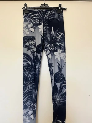Sport Blåa leggings med blommigt mönster - Snygga sport leggings med blått och grått blommigt mönster. De har hög midja och är tillverkade i ett stretchigt material som sitter tight och bekvämt. Perfekta för dig som vill ha en cool och unik look med tropiska vibbar.