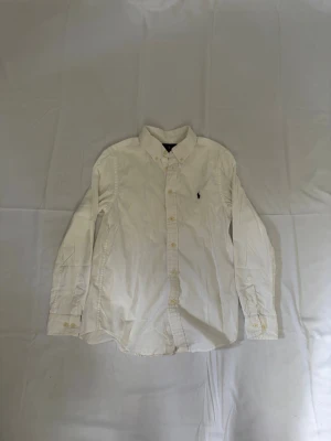 Vit skjorta från Ralph Lauren - Klassisk vit skjorta från Ralph Lauren med button-down krage och broderad logga på bröstet. Skjortan har långa ärmar och knäppning framtill. Perfekt till både jeans och chinos för en clean och stilren look.