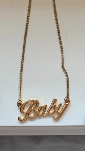 Guldigt halsband med Baby-text - Snyggt halsband i guldfärgad metall med ett hängsmycke där det står 'Baby' i skrivstil. Kedjan är tunn och smidig, perfekt för att ge din outfit en personlig touch. Inget märke syns på halsbandet.