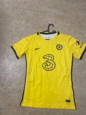 Chelsea FC gul matchtröja Nike - Chelsea FC:s officiella gula matchtröja från Nike med svarta detaljer och klubbmärke på bröstet. Tröjan har korta ärmar, svart logga på ärmen och sponsortryck på framsidan. Tillverkad i lätt och ventilerande material, perfekt för fotbollsträning eller match.