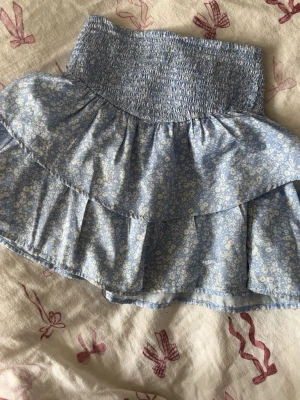 Blommig blå volangkjol med smock - Supersöt kort kjol i ljusblått med vitt blommönster och bred smock i midjan. Kjolen har dubbla volanger som ger en luftig och lekfull känsla. Perfekt för varma dagar och enkel att matcha med en enkel topp.