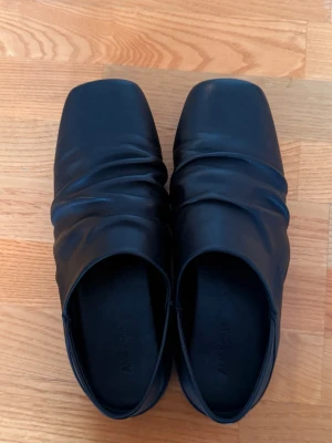 Ambisom mule loafers  - Stilrena svarta loafers från Ambisom i mjukt läder med unik rynkad ovandel och fyrkantig tå. Skorna har en minimalistisk look och passar perfekt till dig som gillar modern och clean stil.
