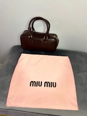 Brun handväska från Miu Miu - Snygg mörkbrun handväska från Miu Miu i slätt läder med guldfärgade metalldetaljer och dubbla handtag. Väskan har en rektangulär form och präglad logotyp på framsidan. Perfekt för dig som vill ha en stilren och trendig accessoar.