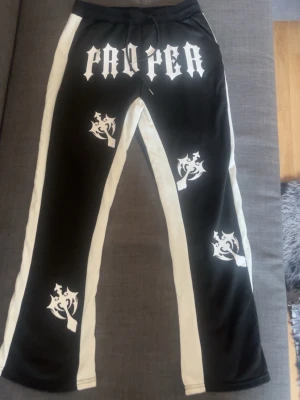 Svarta mjukisbyxor från Proper - Säljer ett par svarta mjukisbyxor från Proper med vita sidor och feta vita tryck på benen och framtill. Byxorna har dragsko i midjan och raka ben. Perfekta för en streetwear-look och riktigt sköna att ha på sig.