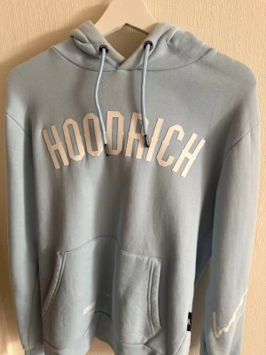 Ljusblå hoodie från Hoodrich - Snygg ljusblå hoodie från Hoodrich med stor vit logga på bröstet och texten 'FROM NOTHING TO SOMETHING' på ryggen. Hoodien har huva med dragsko, magficka och tryck på ena ärmen. Perfekt för en avslappnad och trendig stil.