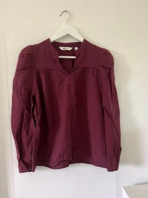 Vinröd blus från Kappahl - Vinröd blus från Kappahl med lång ärm och lätt puff vid axlarna. Blusen har en liten ståkrage med v-ringning och är gjord i ett tunt, mjukt tyg som ger ett avslappnat intryck. Perfekt att styla med jeans eller kjol.