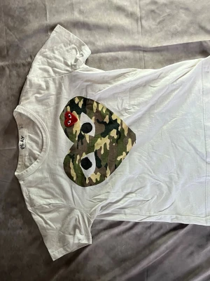 Vit Comme des Garçons PLAY t-shirt - Vit t-shirt från Comme des Garçons PLAY med stort hjärta i camouflage-mönster och en liten röd hjärtdetalj med ögon på bröstet. Klassisk passform och rund hals. Perfekt statement-plagg för dig som gillar streetwear.