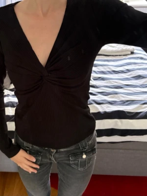 Svart ribbad topp med twist framtill - Svart långärmad topp med djup v-ringning och twistad detalj framtill. Ribbad struktur som ger en snygg och smickrande passform. Perfekt till jeans eller kjol för en trendig look.