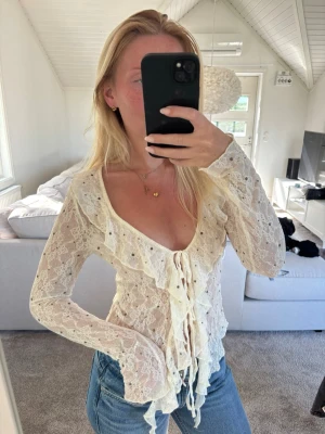 Vit spetsblus med volanger - vit spetsblus med långa ärmar och djup urringning. Blusen har volangdetaljer framtill och knytning i midjan. Tillverkad i genomskinligt spetsmaterial med blommönster
