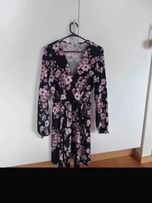 Blommig omlottklänning med knytband - Snygg svart omlottklänning med rosa och vita blommor. Klänningen har v-ringning, långa ärmar och ett knytband i midjan för en smickrande passform. Perfekt för dig som gillar feminina detaljer och blommigt mönster.