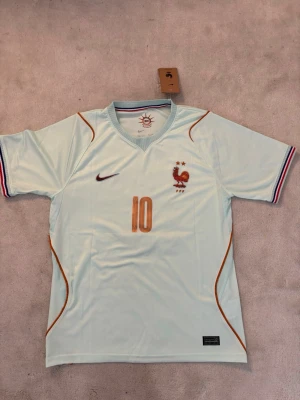 Nike Frankrike fotbollströja ljusblå S och M - Snygg ljusblå fotbollströja från Nike i samarbete med Frankrikes landslag. Tröjan har nummer 10 på bröstet, broderad tupp och två stjärnor, samt FFF-logga. Ränder i blått, vitt och rött på ärmsluten och tunna orange detaljer. Tillverkad i lätt och ventilerande material.