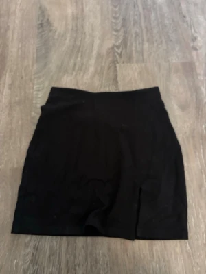 Svart kortkjol med slits - En enkel och stilren svart kortkjol med en liten slits framtill. Kjolen har en rak passform och är perfekt att matcha med olika toppar. Passar dig som gillar minimalistisk stil.