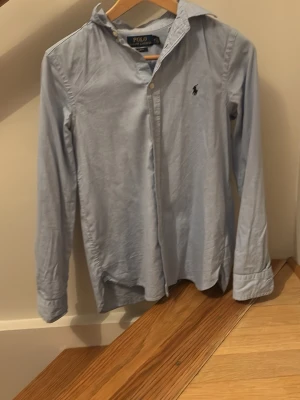 Ljusblå skjorta från Polo Ralph Lauren - Klassisk ljusblå skjorta från Polo Ralph Lauren med broderad logga på bröstet. Skjortan har lång ärm, krage och knappar framtill. Tillverkad i bomull för en skön och stilren look. 
