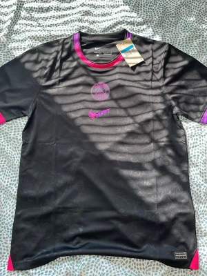 Svart PSG x Nike t-shirt - Snygg svart PSG t-shirt från Nike med lila och rosa detaljer vid hals och ärmslut. Tröjan har PSG-logga och Nike Swoosh framtill i rosa, samt diskret mönster i tyget. Tillverkad i ett lätt och ventilerande material, perfekt för träning eller streetwear.
