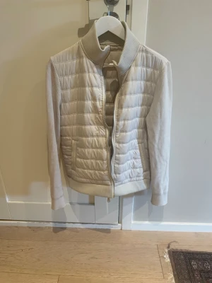 Beige pufferjacka med stickade ärmar - Säljer en beige pufferjacka med quiltat mönster och ribbstickade ärmar samt krage. Jackan har dragkedja framtill, två fickor och en mjuk, bekväm känsla. Perfekt för kalla vinterdagar och ger en clean, stilren look.
