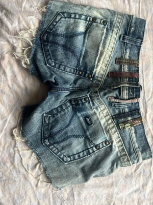 Blå denimshorts med slitningar - Säljer ett par blå denimshorts från Replay med råa, fransiga kanter och slitna detaljer. Shortsen har dekorativa bälteshällor i olika färger och mönster, samt nitar vid fickorna. Klassisk femficksmodell och dragkedja framtill.