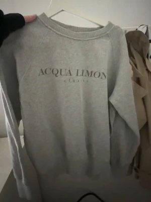 Grå sweatshirt Acqua Limone - Grå sweatshirt från Acqua Limone med tryck framtill och stort citrongult tryck på ryggen. Klassisk rund halsringning och långa ärmar. Perfekt för en avslappnad och trendig stil.