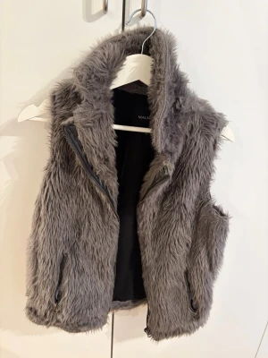 Grå fluffig pälsväst  - Supersnygg grå pälsväst från Malabar i fuskpäls med hög krage och dragkedja framtill. Västen har två fickor med dragkedja och är helfodrad i svart tyg. Perfekt för att addera lite extra stil till din outfit.