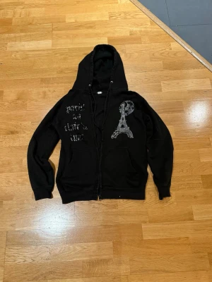 Svart hoodie med strasstryck från Blinglucid - Svart hoodie från Blinglucid med dragkedja, stor huva och fickor framtill. Dekorerad med glittrande strasstenar i form av text och ett Eiffeltorn på bröstet. Tillverkad i mjukt material som känns skönt mot huden. Perfekt för dig som gillar streetstyle och unika detaljer.