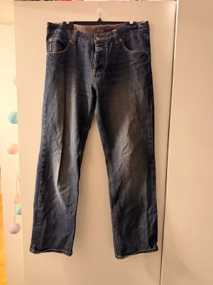 Blå raka jeans med slitningar - Säljer ett par blå jeans med rak passform och snygga slitningar framtill. Jeansen har klassisk femficksdesign, kontrastsömmar och normal midja. Perfekta för en avslappnad och trendig look.