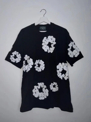 Svart t-shirt med vita blommor - Cool svart t-shirt med stora vita blommor tryckta över hela plagget. T-shirten har rund hals och korta ärmar. Perfekt för dig som vill sticka ut med ett unikt mönster. Materialet känns som bomull och passformen är normal.