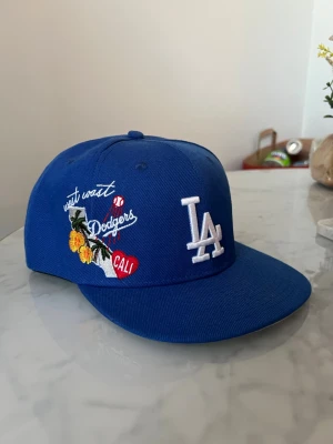 Blå LA Dodgers keps från New Era Storlek 59,6 - Snygg blå keps från New Era med LA Dodgers-logga framtill och broderade detaljer på sidan som palmer, blommor och texten 'west coast' samt 'CALI'. Klassisk rak skärm och broderade ventilationshål. Perfekt för dig som gillar streetwear och sportig stil. Storlek 7 1/2