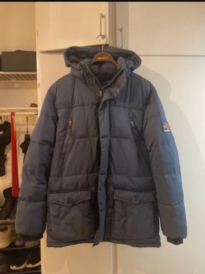 Marinblå Race Marine Dunjacka - Snygg marinblå dunjacka från Race Marine med huva och två stora framfickor med knappar samt två bröstfickor med dragkedja. Jackan är perfekt för kalla vinterdagar. En skavanker som syns på sista bilden. 