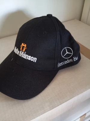 Svart keps Mercedes-Benz & Chrysler - Svart keps med broderade loggor för Mercedes-Benz och Chrysler samt texten 'Matte Månson' i vitt och orange. Klassisk böjd skärm och justerbar passform. Tillverkad i bomullstyg, perfekt för dig som gillar bilar och motorsport.