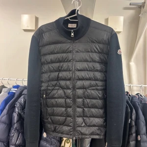 Svart cardiganjacka från Moncler - Snygg svart cardiganjacka från Moncler med quiltad framsida och stickade ärmar. Jackan har dragkedja framtill, ståkrage och två fickor med dragkedja. Moncler-logga på ärmen ger en exklusiv touch. Perfekt för lager-på-lager och streetstyle.
